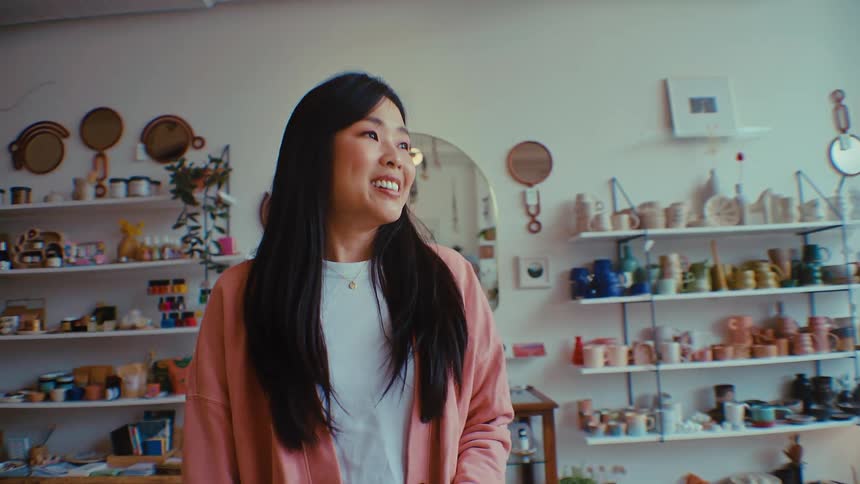 A smiling asian woman wearing a pink cardigan walks through a brightly lit pottery studio and boutique filled with ceramic goods on shelves mirrors and indoor plants
