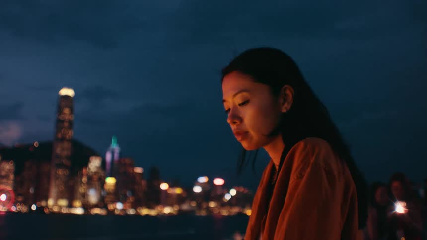 A woman in an orange jacket stands at a waterfront at night looking down thoughtfully before lifting her head to gaze at the illuminated city skyline across the water