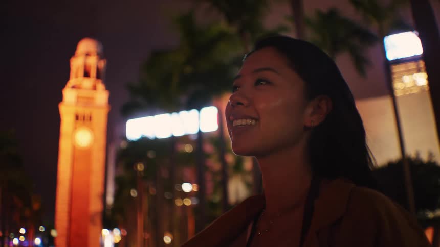 Woman looks at an illuminated nighttime urban scene with a tower and palm trees in the background