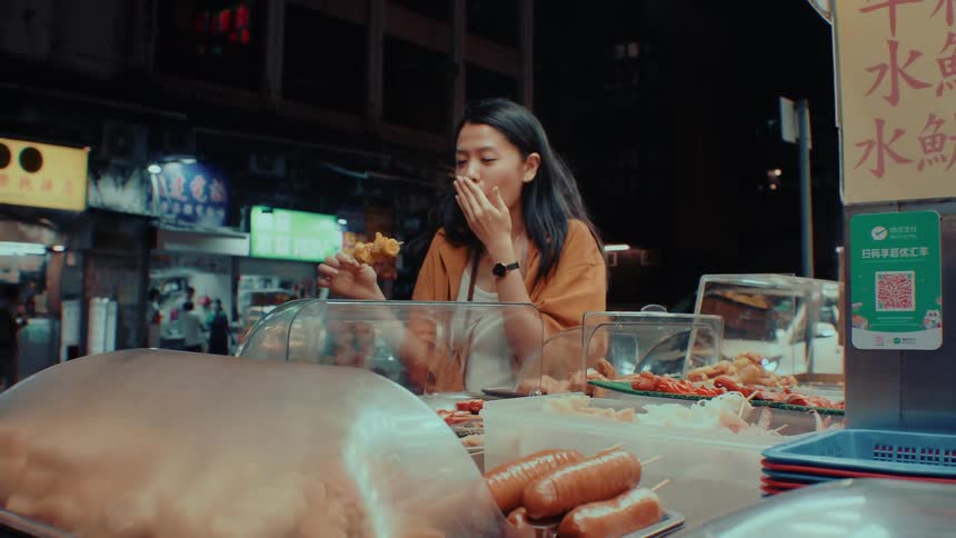 Illuminated by the warm glow of a busy night market stall a young woman takes a bite from a food skewer and covers her mouth while chewing