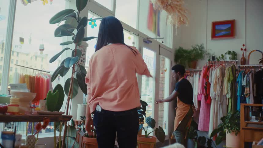 A smiling young woman wearing a pink cardigan stands in a brightly lit boutique filled with plants and clothing where she shares a warm hug with a man wearing an apron before he walks toward the glass door