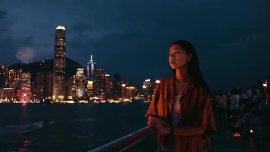 A young woman in a brown shirt leans against a railing at night looking out over the illuminated city skyline and harbor while other people gather in the blurred background