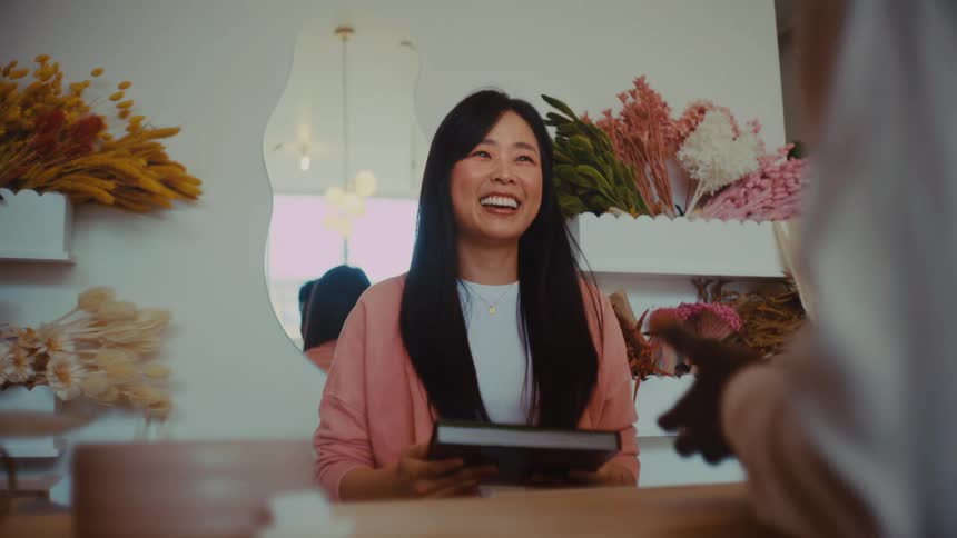 A smiling woman stands behind a wooden counter to receive a black box from a customer engaging in a friendly conversation within a bright floweradorned boutique