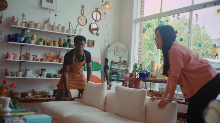 A young black man and a young woman repositioning a couch inside a pottery shop at midday then sharing a warm hug afterward as the woman smiles and thanks him for his help before he heads out