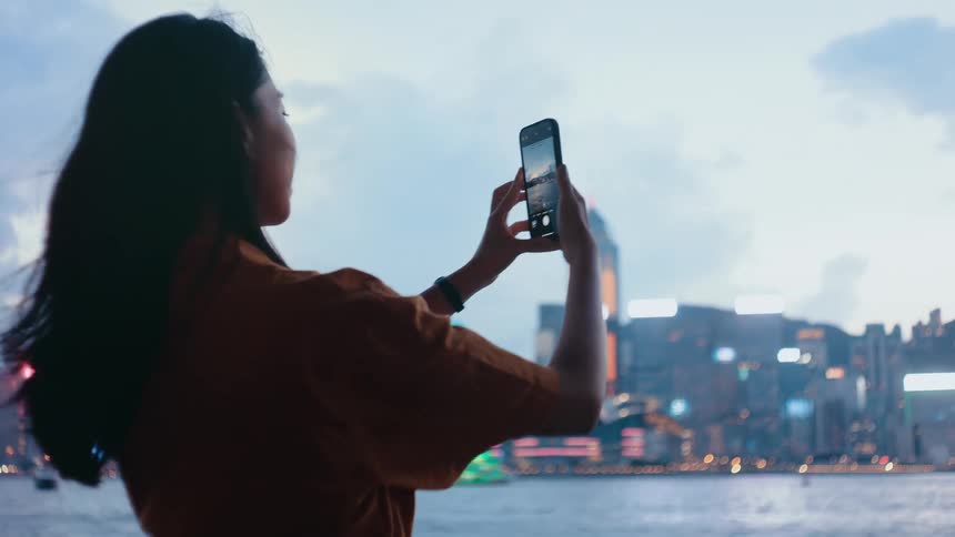 Woman photographing illuminated city skyline at dusk with greenlit boat in harbor