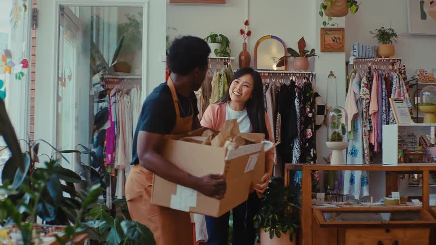 A man and a woman carry cardboard boxes through their colorful clothing boutique while pointing and discussing where to place the new inventory