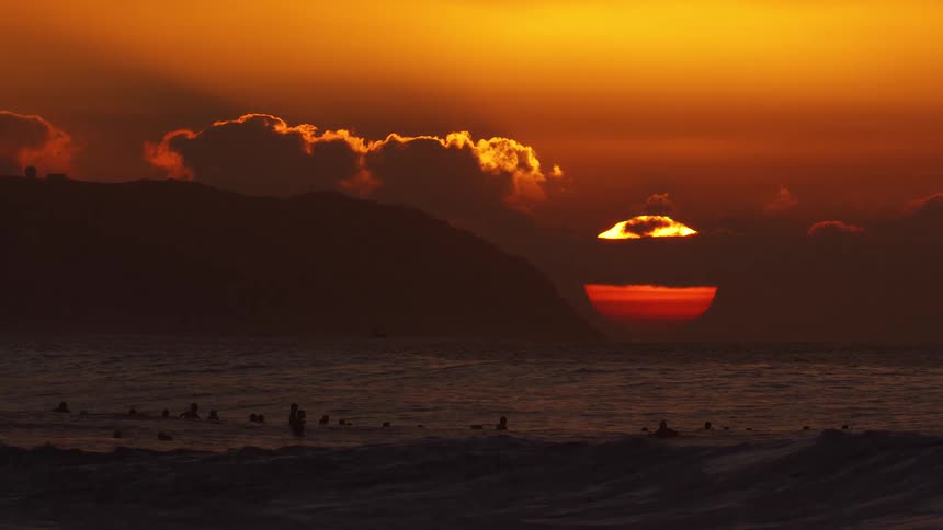 A large deep orange sun sets behind a silhouetted coastline while a group of surfers waits in the dark ocean wavesgolden clouds hang in the glowing sky above the water creating a highcontrast scene of natural beauty and recreation