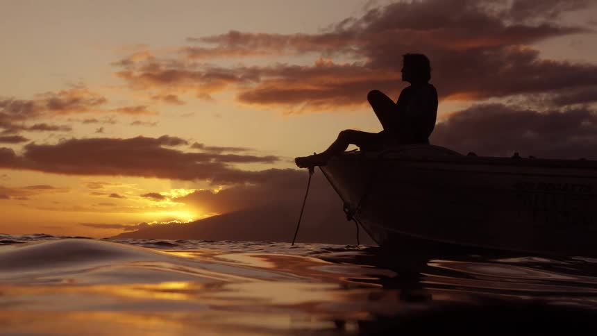 Perched on the bow of a small boat a solitary figure sits in silhouette against a dramatic sunset watching the golden light reflect off the gentle ocean waves
