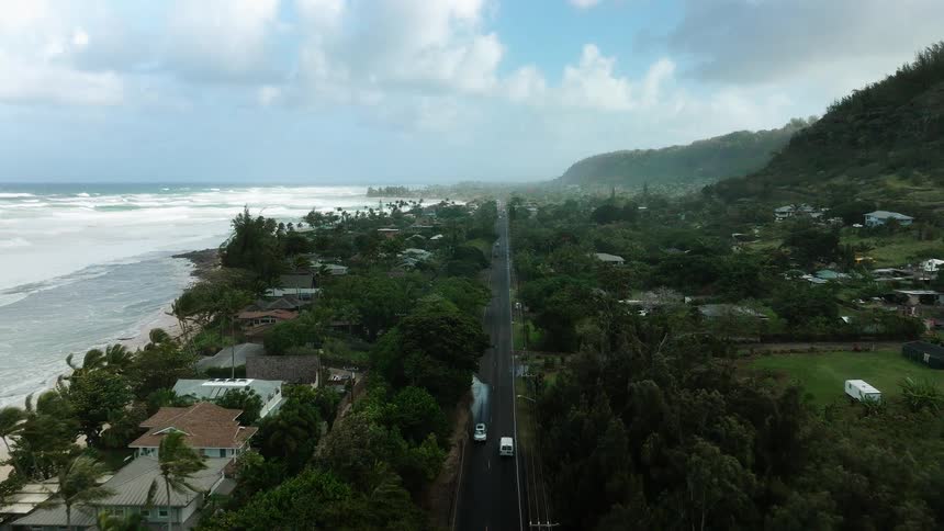 An aerial perspective follows a coastal highway where cars travel between the churning white waves of the ocean and a lush green landscape