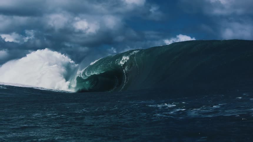 A massive ocean wave swells and curls into a powerful barrel churning up thick white foam and mist against a backdrop of moody clouds and deep blue water