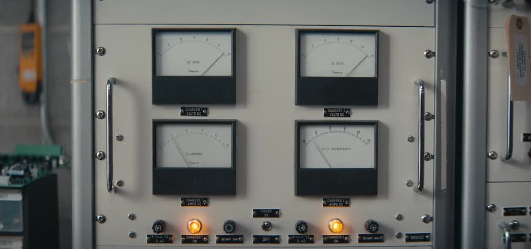 This video features a closeup of a power control rack containing four analog meters that measure dc volts and amperes a hand is shown reaching out to flip a central power switch from the off to the on position causing indicator lights to illuminate