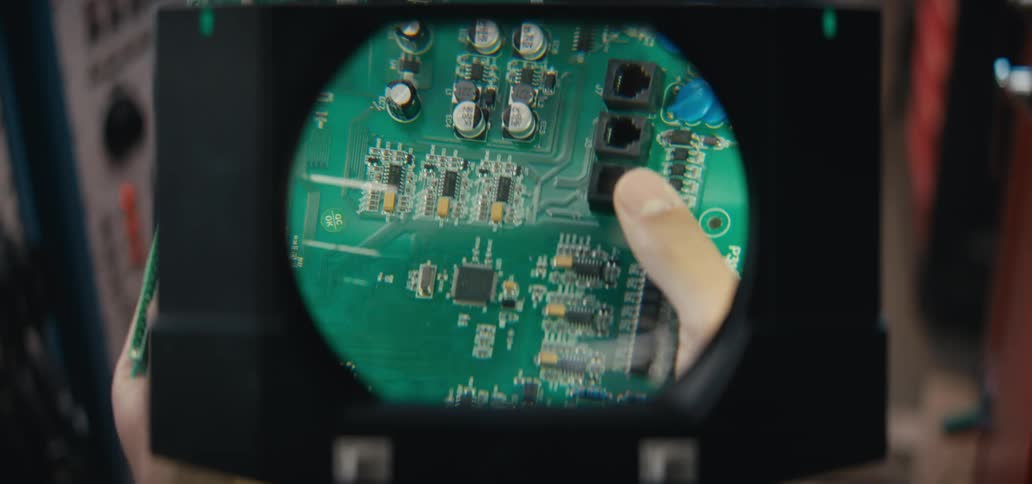 A technician inspects a green printed circuit board through a large circular magnifying lens to check its small components they rotate the board by hand examining various parts like integrated circuits and connection ports under the glass