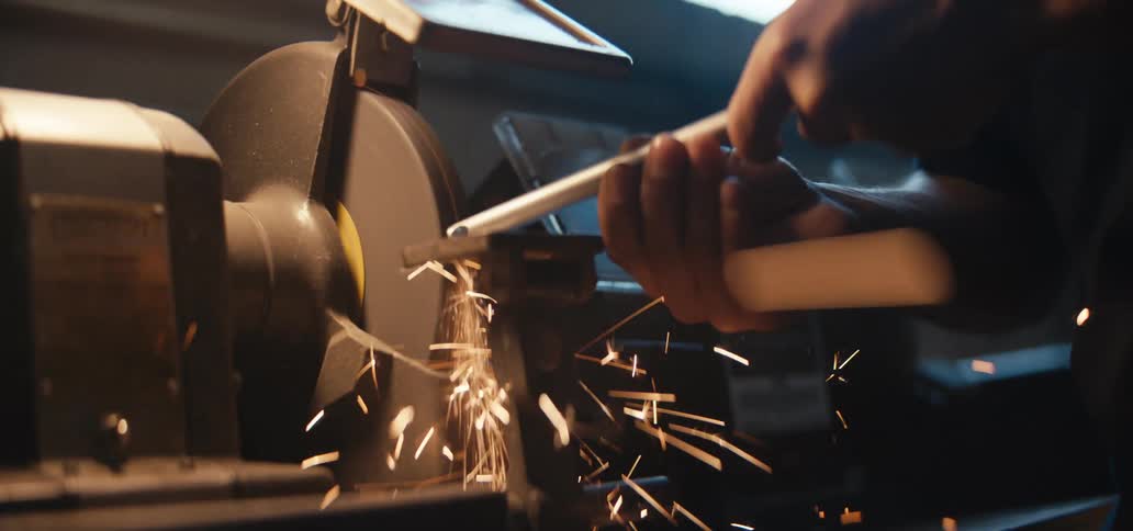 A person sharpens a metal tool on a bench grinder with sparks flying in a workshop