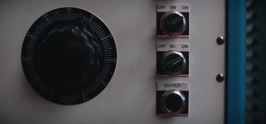 A hand reaches out to turn a silver knob labeled oc from the off position to on the control panel also features a large powerstat dial and several other industrial switches