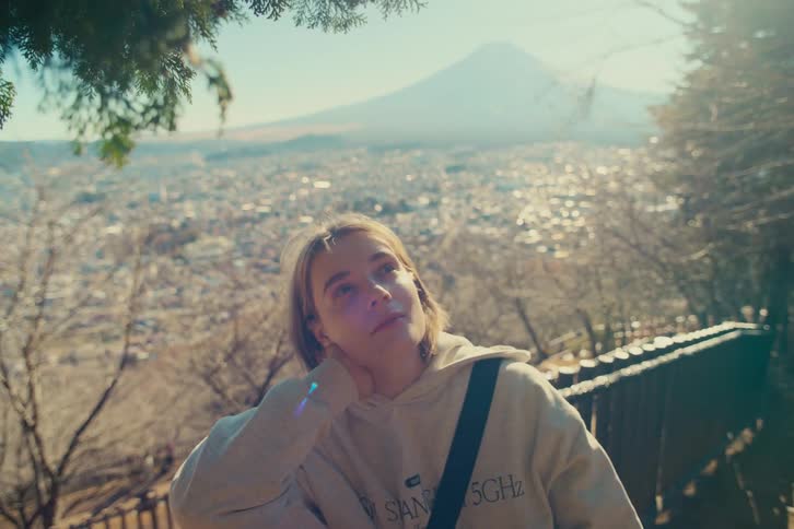 A woman in a beige hoodie sits by a wooden railing and gazes toward a natural scenery and the sprawling city below while leaning her head on her hand during a sunny day