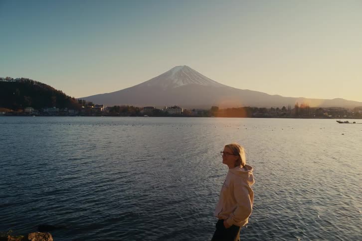 A young woman wearing a yellow jacket and glasses standing on the bank of a lake at sunset holding up his phone to capture a snapshot of the surrounding nature as warm light reflects off the water as a boat drive by