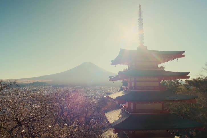 A sunlit multitiered japanese pagoda rises gracefully on a wooded hillside its dark green eaves and red wooden structure sharply silhouetted against a pale blue sky as warm morning light