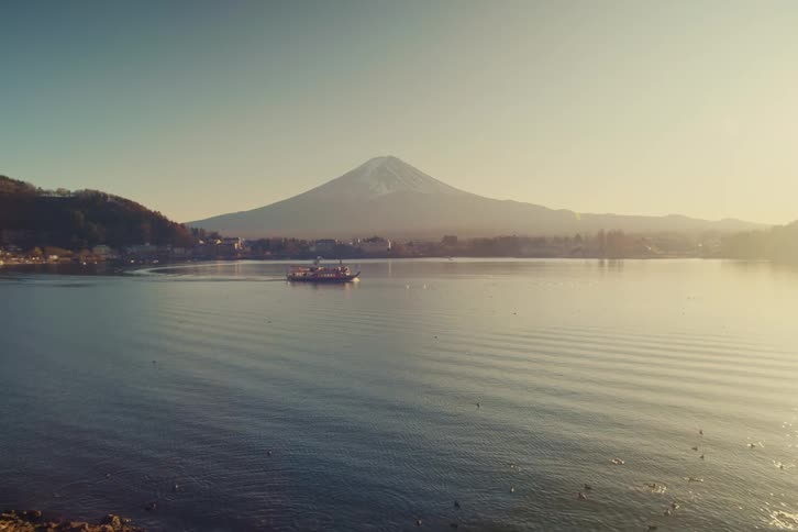 A tour boat glides across the calm shimmering surface of lake kawaguchi while the snowcapped peak of mount fuji stands majestically under the soft golden light of a morning sky