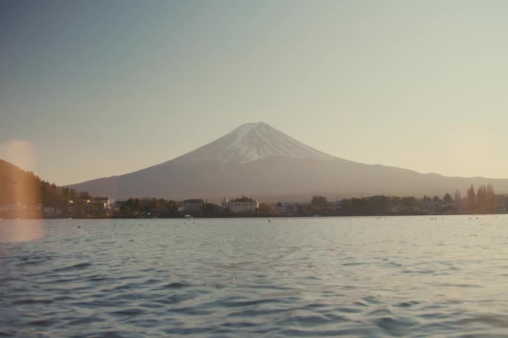 A lake with windstirred water swaying gently a small town resting along the treelined bank and a volcanic mountain rising in the background as the sun sets behind it