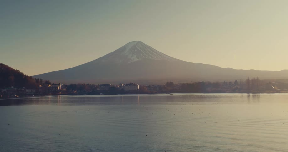 A wide static shot shows mount fuji with a snowcovered summit towering over a lakeside town during a clear morning