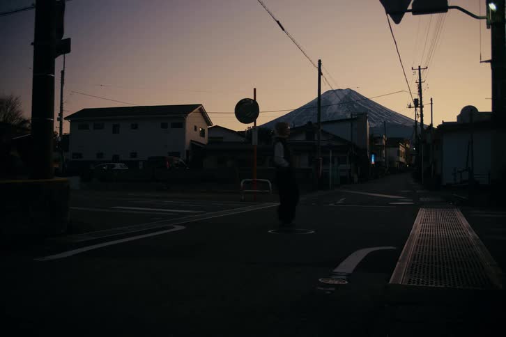 Woman skips slightly while walking through an intersection in a residential neighborhood while red tints the sky around a mountain in the distance