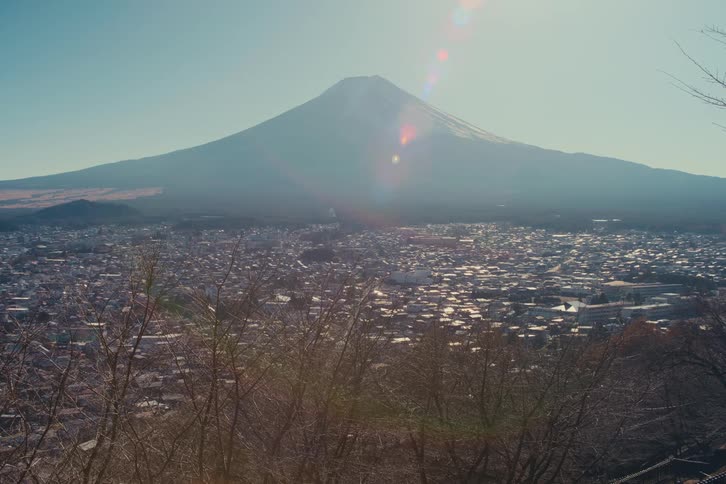 A static highangle view of a vulcanic mountain towering over a sprawling city under a bright sun with leafless tree branches framing the bottom of the shot while a lens flare appears near the mountain peak