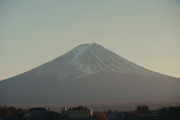 A mountain with a town in the foreground its peak covered in snow as the sun sets behind it