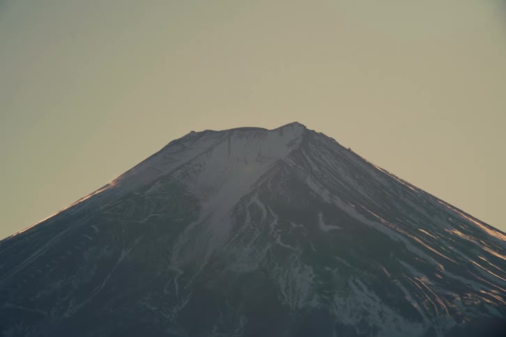 The snowcovered peak of mount fuji stands prominently against a pale evening sky its rugged ridges and volcanic crater illuminated by the soft warm glow of a setting sun