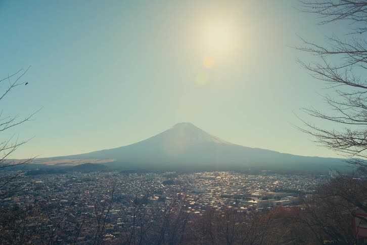 A vast city spreads across the valley beneath a clear winter sky with mount fujis snowdusted perfectly conical peak rising serenely in the distance as bare tree branches frame the view sunlight glinting off rooftops and casting a warm haze