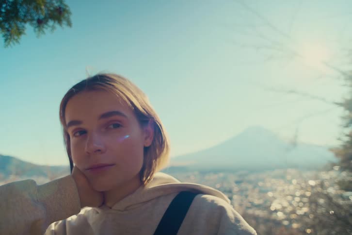 A young woman rests her chin on her hand at a scenic outlook looking toward the camera and then away as golden sunlight and a soft lens flare dance around her with the hazy silhouette of mount fuji visible behind her