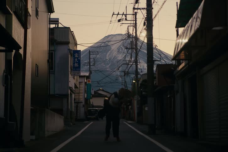 At the far end of the quiet street the man gently lifts the woman onto his back and carries her forward beneath the tangle of power lines their figures silhouetted against the towering snowcapped mountain in the fading golden light