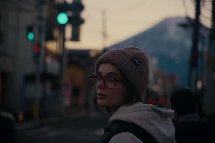 Woman looks around while standing next to an urban road leading towards a snowy mountain