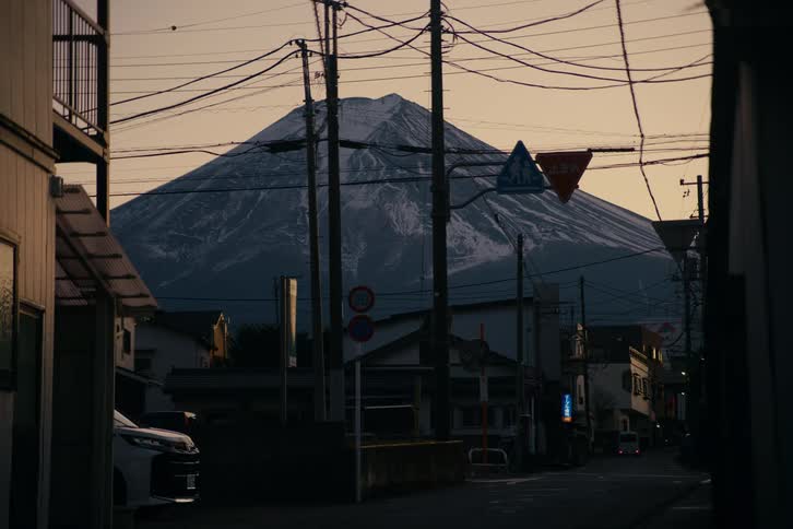At dusk a quiet residential street lined with modest houses and parked cars leads the eye toward mount fuji looming dramatically in the background its snowstreaked slopes rising behind a dense web of power lines and street signs