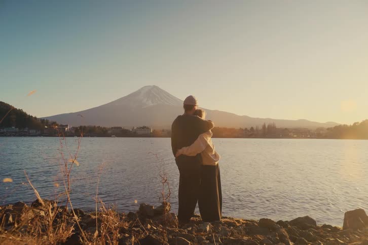 A couple standing on a riverbank hugging each other as they look toward a mountain on the opposite side during a warm glowing sunset