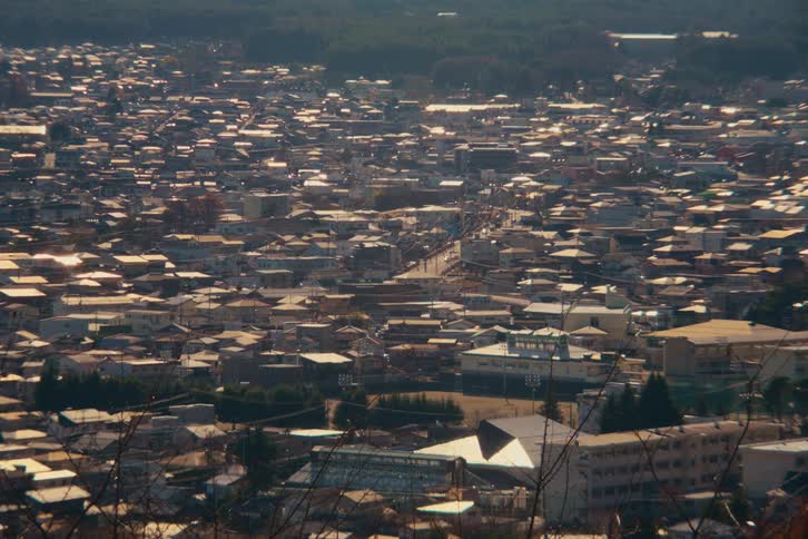 A highangle wide shot shows a dense residential neighborhood packed with houses and small buildings under a bright morning sun