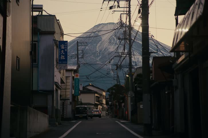 Buildings on a street as a car passes by a parked truck with a mountain in the distance