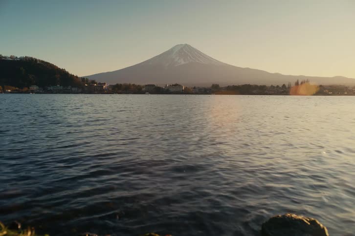 Mount fuji stands out in the background beyond a lake with a rippled surface and a rocky shoreline in the foreground under soft light