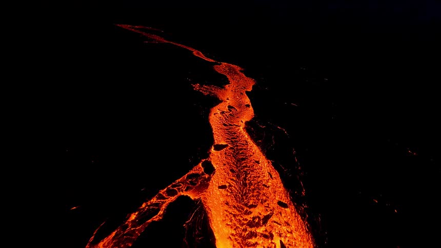 An aerial perspective captures a river of glowing orange lava snaking across a blackened volcanic landscape at night its molten surface rippling with intense heat as it illuminates the surrounding darkness with a fiery light