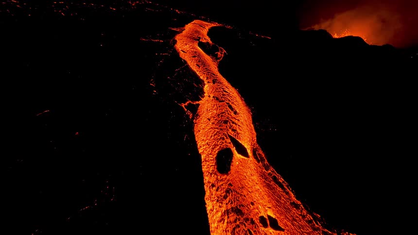 A glowing river of molten lava snakes through the darkness its fiery orange surface bubbling and cracking as it slowly advances casting an intense otherworldly light against the surrounding black volcanic landscape