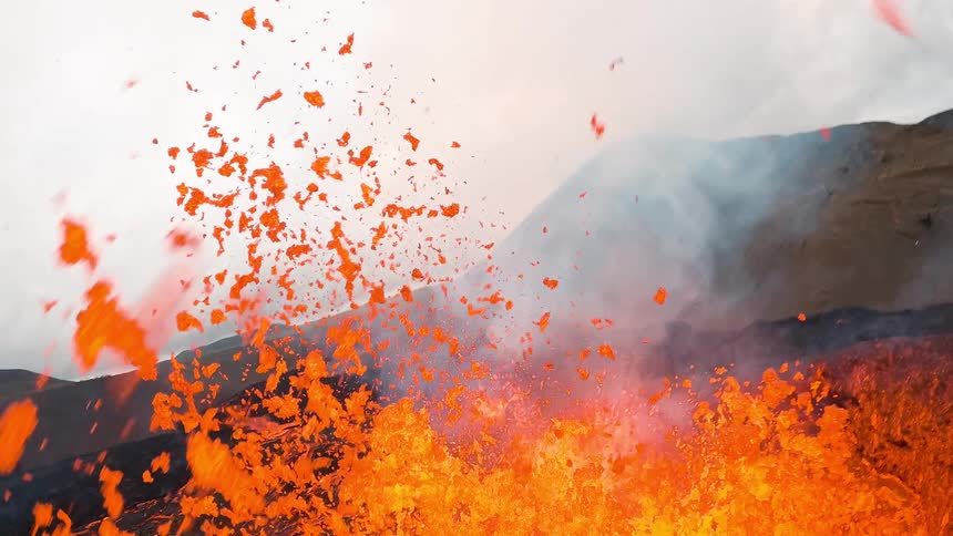 A drone flies over an active volcano as bright orange lava fountains erupt from a vent splattering molten rock across a dark cooling crust while smoke and steam rise against a cloudy sky