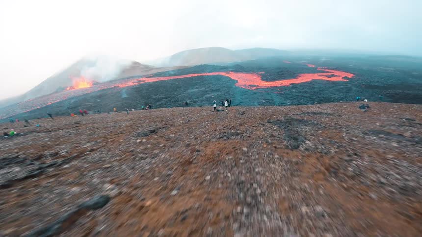A fastmoving drone sweeps down a rocky mountain slope soaring past scattered onlookers before flying low over a vast smoldering lava crust toward a vibrant river of glowing molten rock and a distant erupting volcano