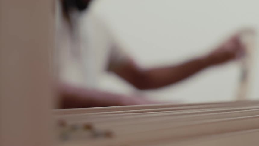 The video shows a closeup of a wooden loom with white threads being meticulously woven by a person in the backgroundthe camera focuses on the intricate movement of the looms components as the artisan works to create a textile
