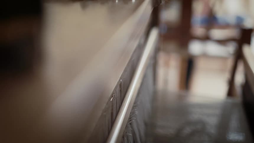 A hand reaches along the length of a wooden loom guiding and adjusting the taut vertical threads beside a smooth rail