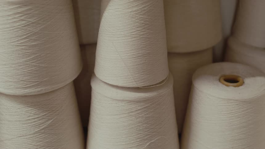 Several large cones of creamcolored yarn are stacked closely together filling the frame the camera remains still as it captures the fine textured threads wound around the conical spools