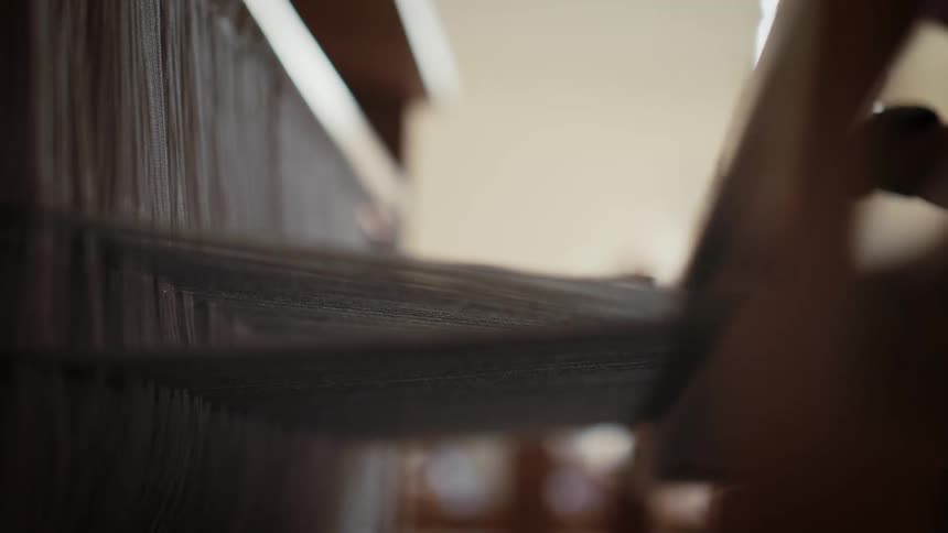 Closeup of a hand weaving dark fabric on a mechanical loom in a factory