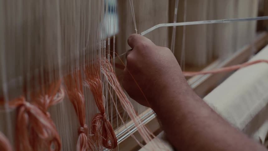 A closeup shot shows a persons hands using a thin metal tool to meticulously thread orange and white yarns through the vertical warp threads of a traditional wooden loom to set up a textile pattern