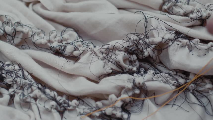 A softly lit expanse of pale fabric rests in gentle folds densely stitched with uneven clusters of white knots and loose black threads while a hand moves across the surface as it carefully sews
