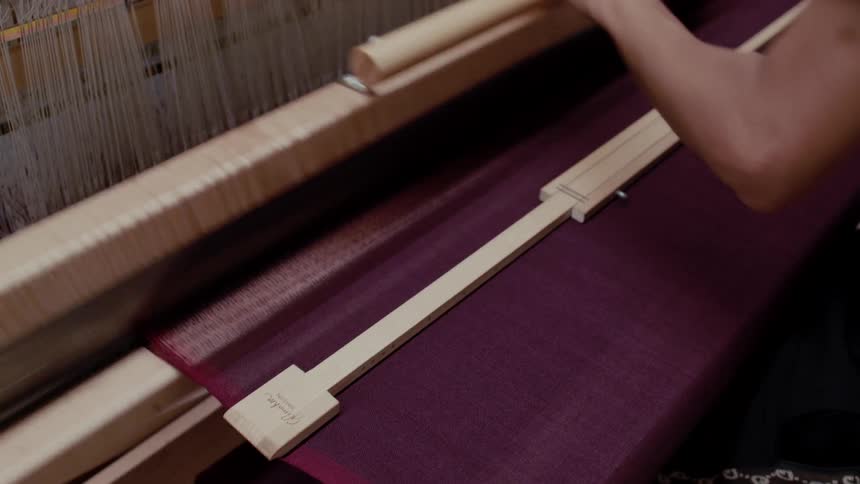 A weaver operates a traditional wooden loom with rhythmic precision skillfully passing a shuttle through the delicate white warp threads and pulling the reed forward to pack the dark maroon fabric into place