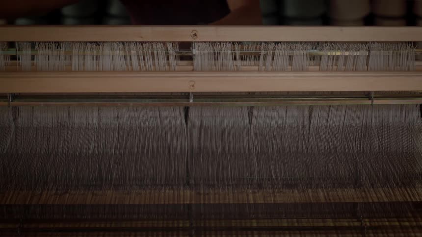Fine white threads stretch taut across a traditional wooden loom as a craftsmans hands move rhythmically in the background to adjust the complex mechanism all captured in the warm quiet atmosphere of a weaving studio