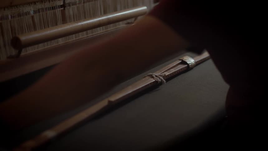A hand reaches along the length of a wooden loom guiding and adjusting the taut vertical threads beside a smooth rail
